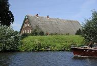 011_26063 Ein strohgedecktes Bauernhaus hinter dem Elbdeich der Dove-Elbe; das Strohdach des Hauses ragt hinter dem Deich hervor auf dem ein Radrennfahrer mit seinem Fahrrad fhrt. -auf dem Wasser der Dove-Elbe kommt ein holzgeklinkertes Motorboot, dessen Bug und Reling schon zu erkennnen ist.     www.christoph-bellin.de